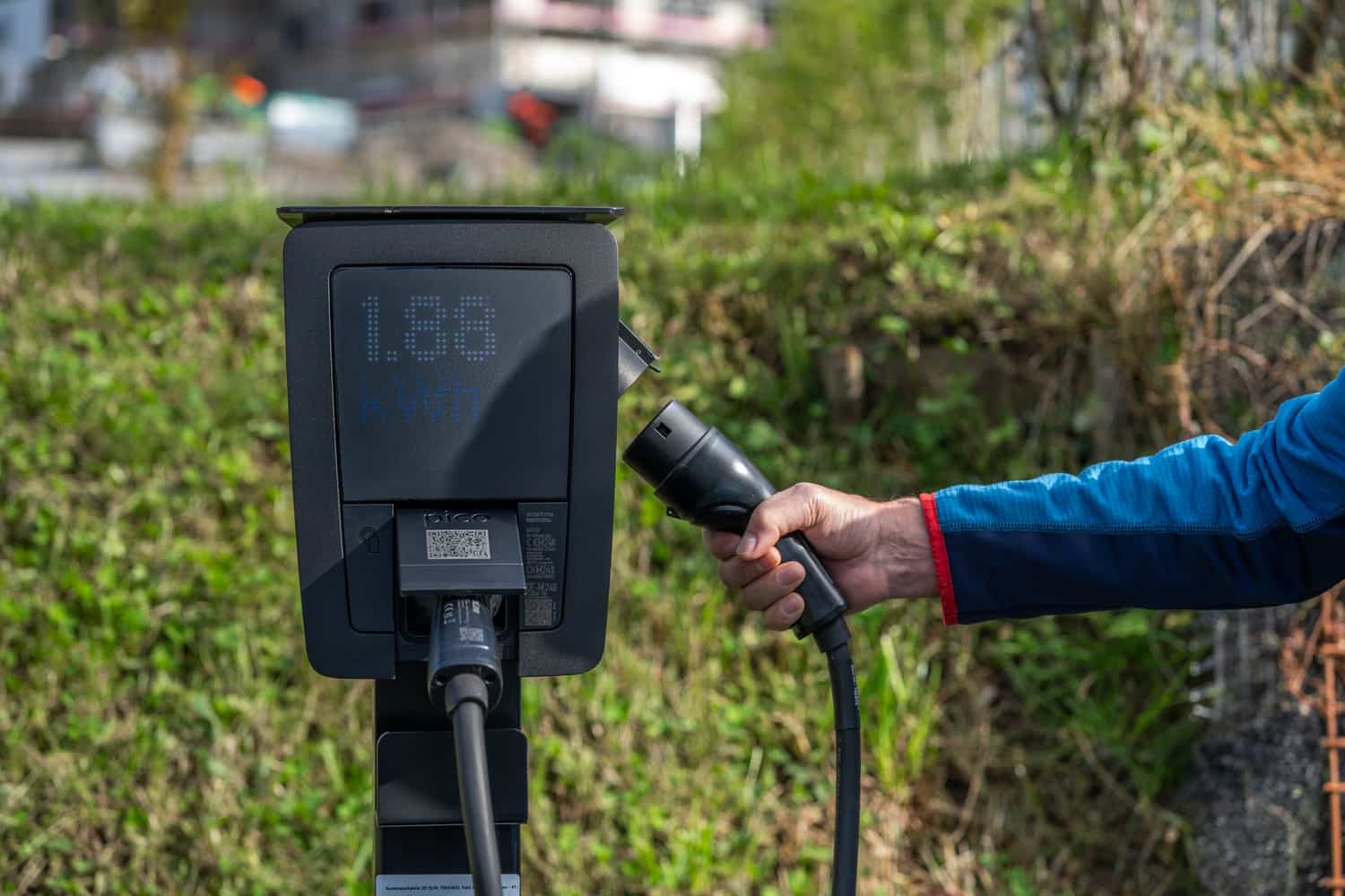 A hand holding a charging connector at an outdoor charging unit showing a low energy reading
